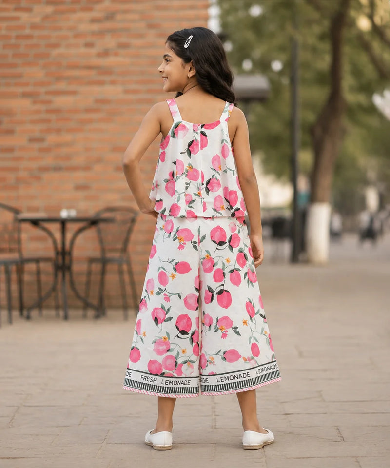 A young girl is shown from behind wearing a white sleeveless jumpsuit with a pink and green watercolor floral print. The jumpsuit features ruffled straps and a wide leg, with a black and white patterned hem that reads 'LEMONADE'.