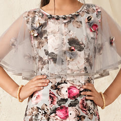 A woman wearing a sheer tulle dress with a large floral print in pink, gray, and black on a light background, accessorized with gold bracelets.