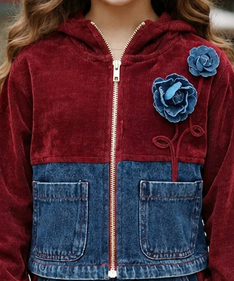 A close-up of a child wearing a zip-up hooded jacket made from a burgundy velvet fabric and blue denim. The jacket features two large blue fabric flowers on the chest, a gold zipper, and two front patch pockets.