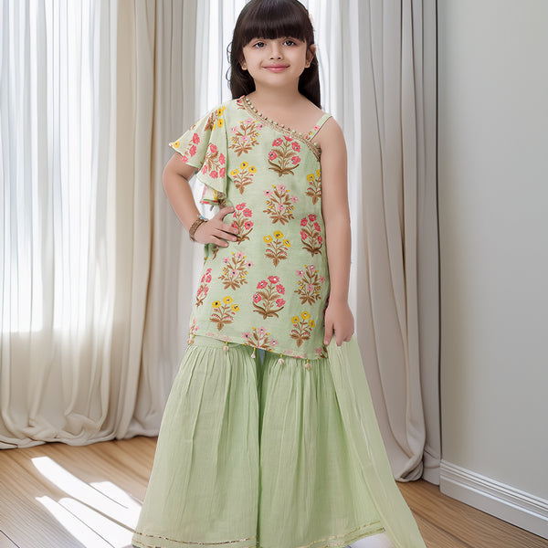 Young girl wearing a floral top and green skirt standing in a room with white curtains.