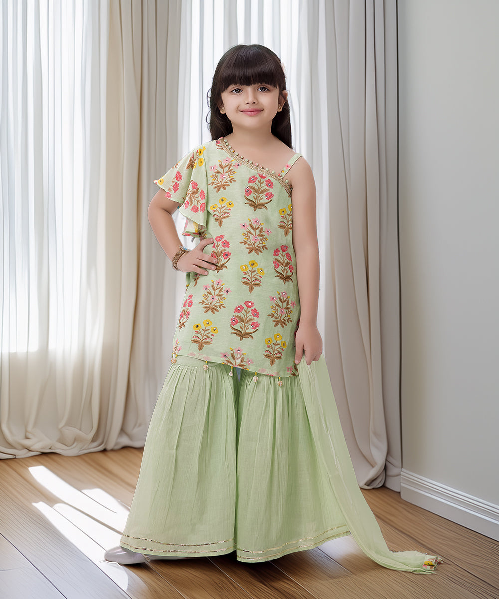 Young girl wearing a floral top and green skirt standing in a room with white curtains.