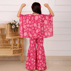 Child wearing a pink floral outfit with arms raised in a room with a wicker table and flowers.