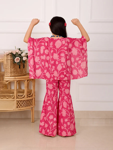 Child wearing a pink floral outfit with arms raised in a room with a wicker table and flowers.