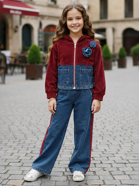 A young girl stands in a cobblestone outdoor plaza wearing a two-tone zip-up jacket made of burgundy velvet and blue denim, paired with wide-leg blue jeans featuring red side stripes and white sneakers.