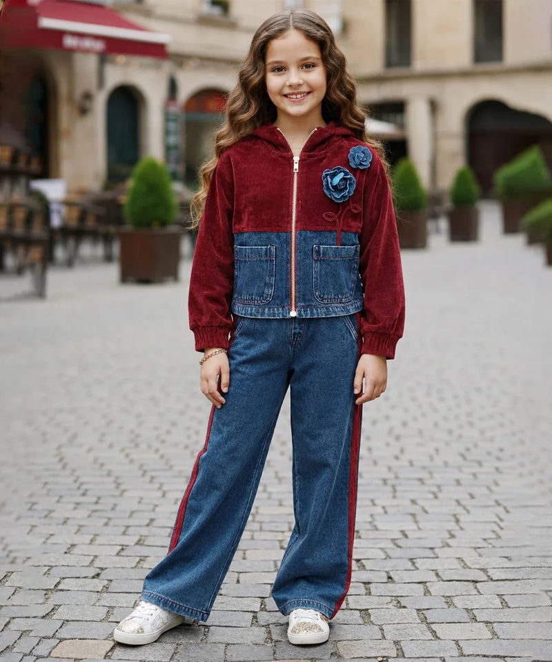A young girl stands in a cobblestone outdoor plaza wearing a two-tone zip-up jacket made of burgundy velvet and blue denim, paired with wide-leg blue jeans featuring red side stripes and white sneakers.