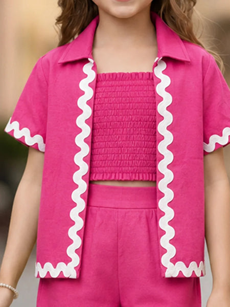 A child wearing a bright pink matching outfit with a short-sleeve collared shirt featuring white wavy trim, a smocked crop top, and matching shorts.