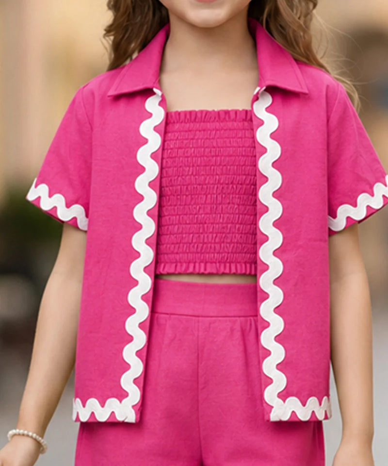 A child wearing a bright pink matching outfit with a short-sleeve collared shirt featuring white wavy trim, a smocked crop top, and matching shorts.