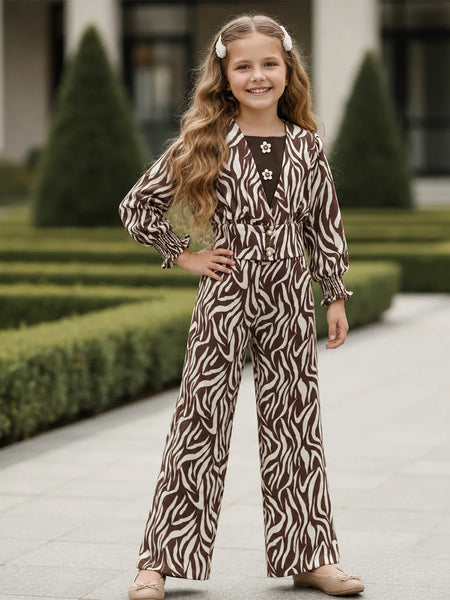 A young girl with long wavy light brown hair stands outdoors on a paved walkway wearing a brown and beige zebra-print jumpsuit with a tie waist and matching cardigan. She is posing with one hand on her hip, with manicured nails and beige shoes visible.