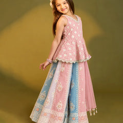 A young girl modeling a traditional Indian outfit consisting of a pink sleeveless kurta with white embroidery and a matching wide skirt in pink and light blue with intricate floral patterns and borders, set against a solid olive green studio background.