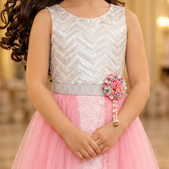 A young girl wearing a sleeveless silver sequined top and a pink tulle skirt dress, with a decorative floral waist accessory, photographed indoors against a softly blurred warm background.