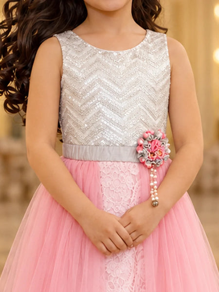 A young girl wearing a sleeveless silver sequined top and a pink tulle skirt dress, with a decorative floral waist accessory, photographed indoors against a softly blurred warm background.