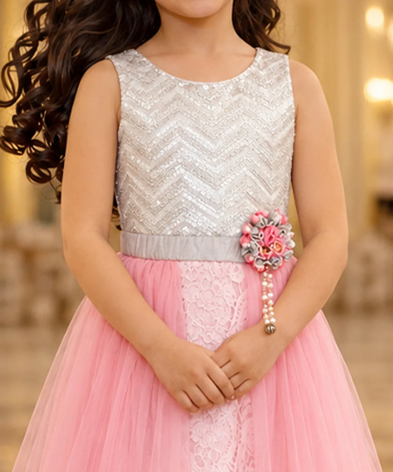 A young girl wearing a sleeveless silver sequined top and a pink tulle skirt dress, with a decorative floral waist accessory, photographed indoors against a softly blurred warm background.