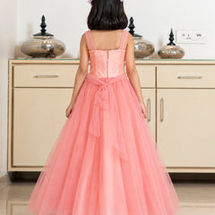 A young girl viewed from behind wearing a coral pink formal dress with a structured corset-style bodice, thin straps, and a full layered tulle skirt, standing in front of a cream-colored cabinet.