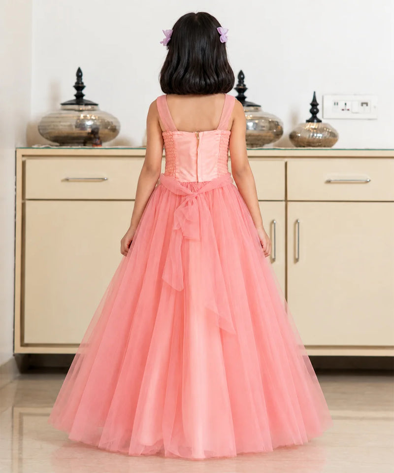 A young girl viewed from behind wearing a coral pink formal dress with a structured corset-style bodice, thin straps, and a full layered tulle skirt, standing in front of a cream-colored cabinet.