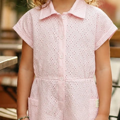 A child wearing a light pink short-sleeve eyelet romper with a button front and collar, photographed in a softly blurred outdoor setting.