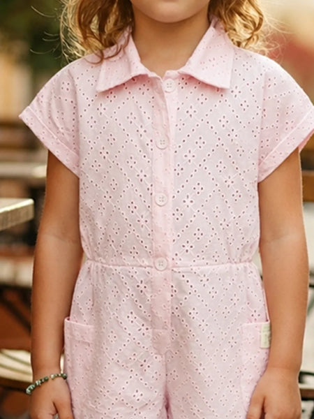 A child wearing a light pink short-sleeve eyelet romper with a button front and collar, photographed in a softly blurred outdoor setting.