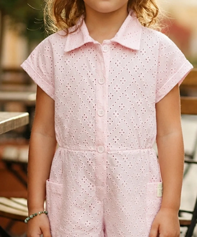 A child wearing a light pink short-sleeve eyelet romper with a button front and collar, photographed in a softly blurred outdoor setting.