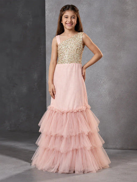 A young girl models a formal one-shoulder mermaid gown with a gold sequined bodice and a layered pink tulle skirt, set against a textured gray studio backdrop.