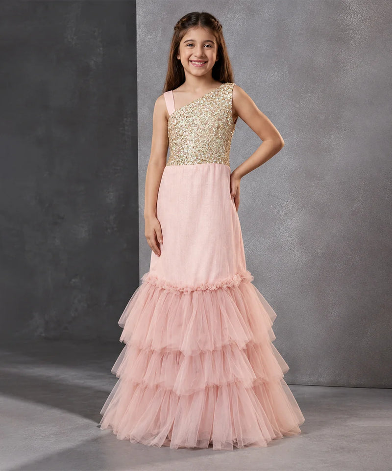 A young girl models a formal one-shoulder mermaid gown with a gold sequined bodice and a layered pink tulle skirt, set against a textured gray studio backdrop.
