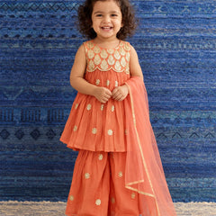 A young girl with curly dark hair stands smiling in a traditional Indian outfit. The outfit consists of a coral orange sleeveless kurta with gold embroidery, matching wide-leg pants, and a sheer orange dupatta draped over one shoulder.