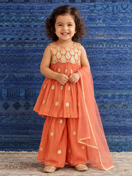 A young girl with curly dark hair stands smiling in a traditional Indian outfit. The outfit consists of a coral orange sleeveless kurta with gold embroidery, matching wide-leg pants, and a sheer orange dupatta draped over one shoulder.