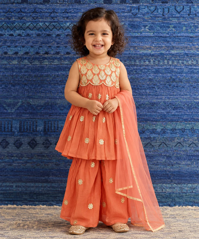 A young girl with curly dark hair stands smiling in a traditional Indian outfit. The outfit consists of a coral orange sleeveless kurta with gold embroidery, matching wide-leg pants, and a sheer orange dupatta draped over one shoulder.