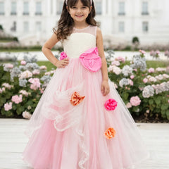 A young girl stands outdoors in front of a large white building and flower beds, wearing a formal ball gown with a cream bodice, bright pink skirt, large pink bow, and pink and orange fabric flowers.