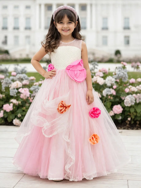 A young girl stands outdoors in front of a large white building and flower beds, wearing a formal ball gown with a cream bodice, bright pink skirt, large pink bow, and pink and orange fabric flowers.