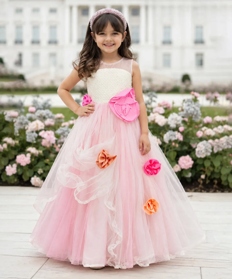 A young girl stands outdoors in front of a large white building and flower beds, wearing a formal ball gown with a cream bodice, bright pink skirt, large pink bow, and pink and orange fabric flowers.