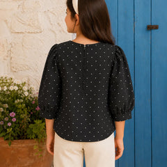 A woman shown from the back wearing a black three-quarter sleeve blouse with white polka dots and puffed shoulders, paired with cream pants and a white headband, standing in front of a blue wooden door and a white stucco wall.