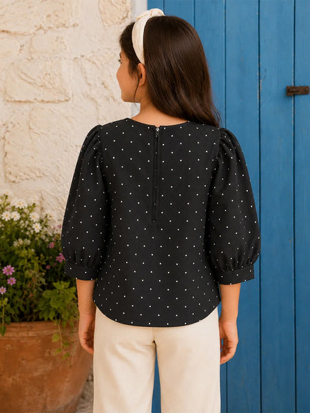 A woman shown from the back wearing a black three-quarter sleeve blouse with white polka dots and puffed shoulders, paired with cream pants and a white headband, standing in front of a blue wooden door and a white stucco wall.