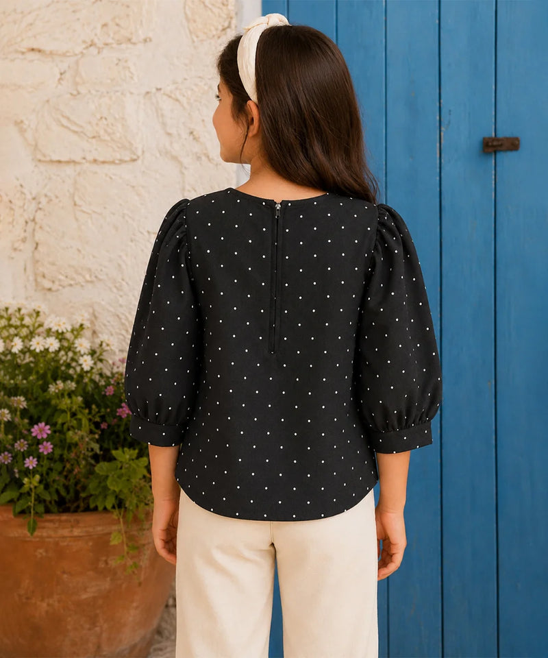 A woman shown from the back wearing a black three-quarter sleeve blouse with white polka dots and puffed shoulders, paired with cream pants and a white headband, standing in front of a blue wooden door and a white stucco wall.