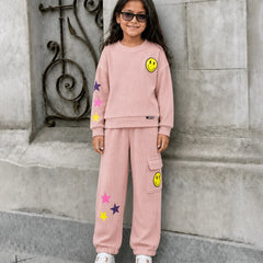 A young girl with long dark hair and sunglasses stands outdoors against a gray stone wall, wearing a light pink matching sweatshirt and jogger pants set with yellow smiley face and star patches.