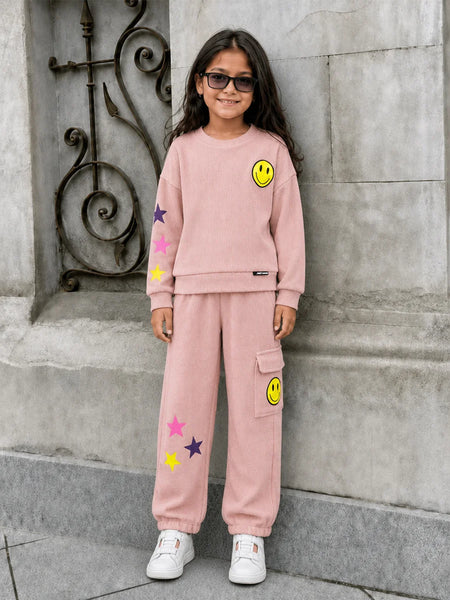 A young girl with long dark hair and sunglasses stands outdoors against a gray stone wall, wearing a light pink matching sweatshirt and jogger pants set with yellow smiley face and star patches.