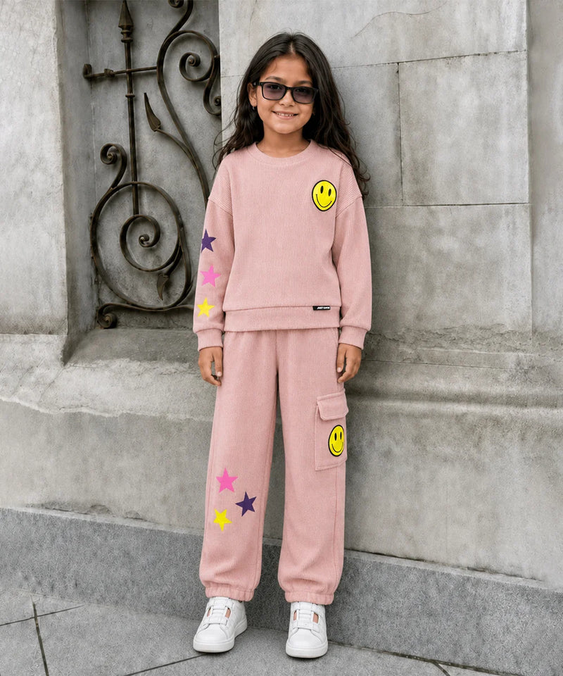 A young girl with long dark hair and sunglasses stands outdoors against a gray stone wall, wearing a light pink matching sweatshirt and jogger pants set with yellow smiley face and star patches.