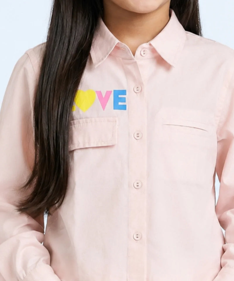 A close-up of a model wearing a light pink button-down shirt with a colorful 'LOVE' graphic on the chest, featuring a yellow heart and blue and pink letters.