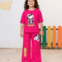 A young girl stands smiling in a two-piece bright pink Snoopy pajama set. The short-sleeve t-shirt features a large Snoopy graphic with 'SNOOPY' printed below, and the matching wide-leg pants have a Snoopy graphic on the thigh and 'SNOOPY' printed vertically down one leg.
