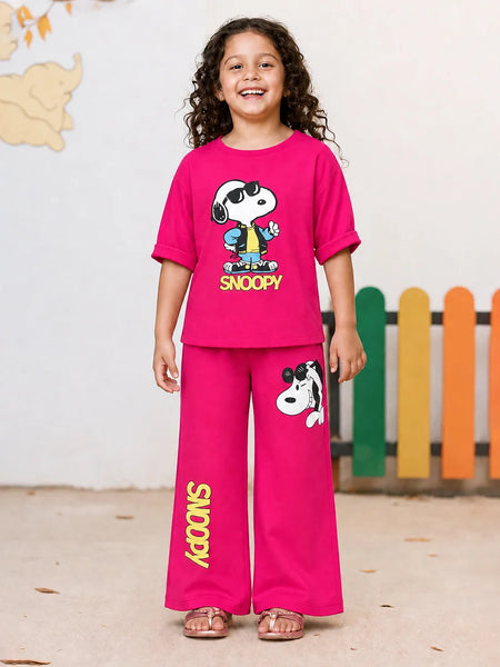 A young girl stands smiling in a two-piece bright pink Snoopy pajama set. The short-sleeve t-shirt features a large Snoopy graphic with 'SNOOPY' printed below, and the matching wide-leg pants have a Snoopy graphic on the thigh and 'SNOOPY' printed vertically down one leg.