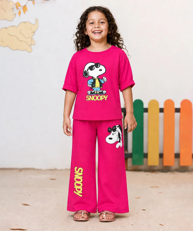 A young girl stands smiling in a two-piece bright pink Snoopy pajama set. The short-sleeve t-shirt features a large Snoopy graphic with 'SNOOPY' printed below, and the matching wide-leg pants have a Snoopy graphic on the thigh and 'SNOOPY' printed vertically down one leg.