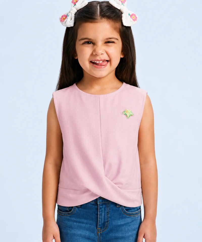A young girl with a white bunny headband smiles while wearing a sleeveless light pink top with a twisted front detail and a small green and yellow star pin, paired with blue jeans against a light blue background.