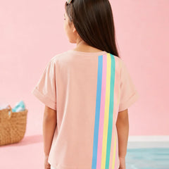 A child model shown from behind wearing a light pink short-sleeve t-shirt with three vertical rainbow stripes (red, orange, yellow, green, blue, purple) on the back, set against a pastel pink background.