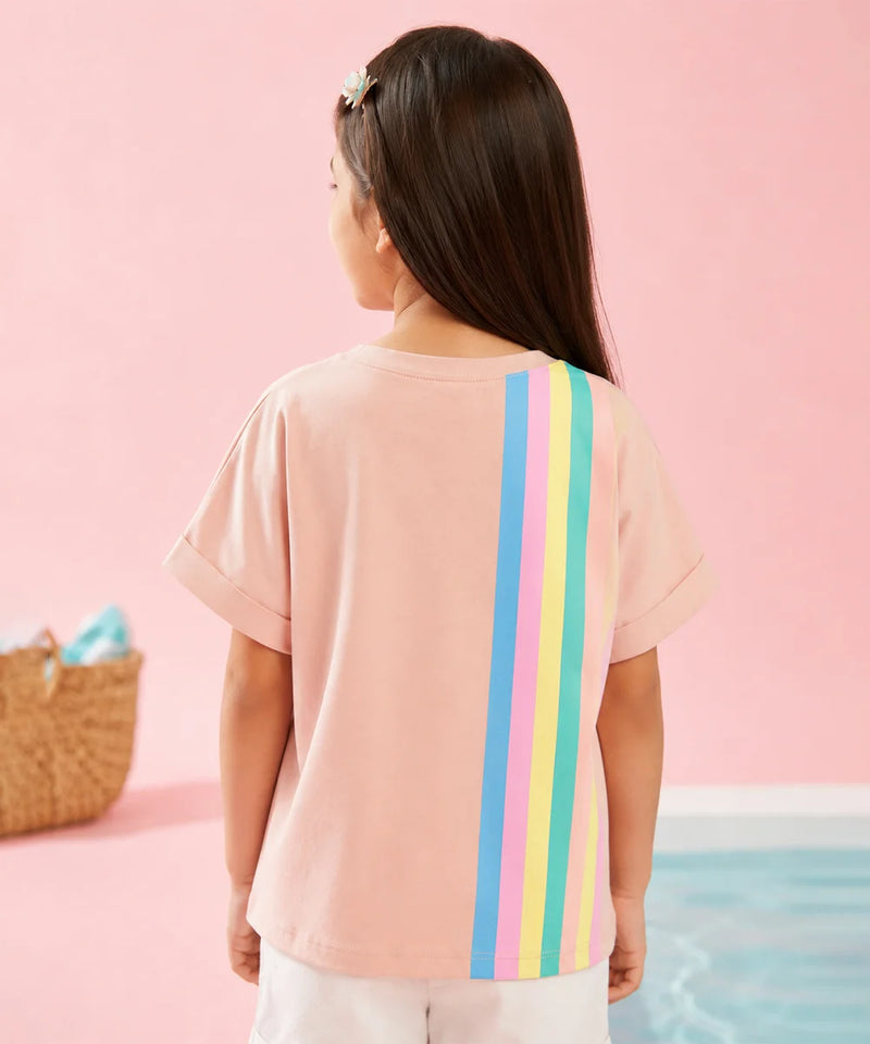 A child model shown from behind wearing a light pink short-sleeve t-shirt with three vertical rainbow stripes (red, orange, yellow, green, blue, purple) on the back, set against a pastel pink background.