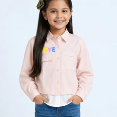 A young girl with long dark hair wearing a light pink button-up shirt with a colorful 'LOVE' graphic on the chest, paired with blue jeans, standing against a light blue background.