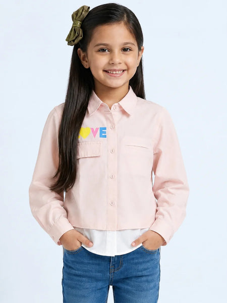 A young girl with long dark hair wearing a light pink button-up shirt with a colorful 'LOVE' graphic on the chest, paired with blue jeans, standing against a light blue background.