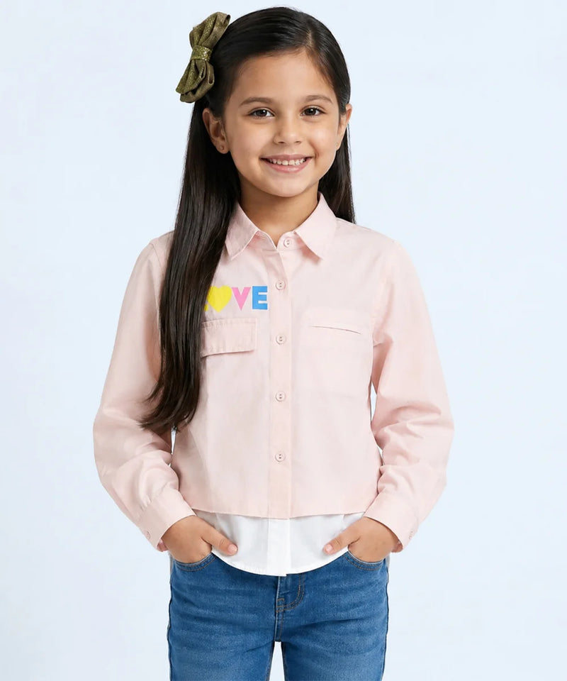 A young girl with long dark hair wearing a light pink button-up shirt with a colorful 'LOVE' graphic on the chest, paired with blue jeans, standing against a light blue background.