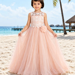 A young girl stands on a sandy beach wearing a sleeveless blush pink ball gown with a lace bodice, floral applique, and a large flower detail at the waist. The background shows a tropical beach scene with palm trees, ocean, and sky.