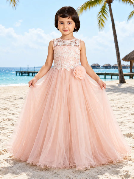 A young girl stands on a sandy beach wearing a sleeveless blush pink ball gown with a lace bodice, floral applique, and a large flower detail at the waist. The background shows a tropical beach scene with palm trees, ocean, and sky.
