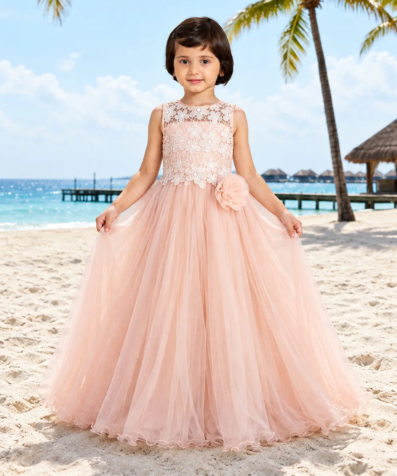 A young girl stands on a sandy beach wearing a sleeveless blush pink ball gown with a lace bodice, floral applique, and a large flower detail at the waist. The background shows a tropical beach scene with palm trees, ocean, and sky.