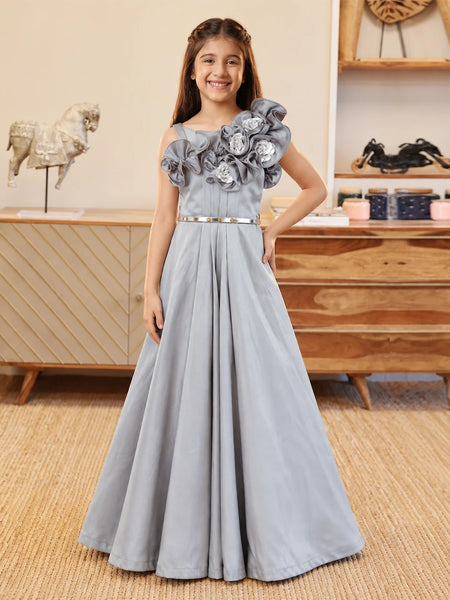 A young girl stands in a room wearing a formal light blue-gray gown with large floral appliqué on the bodice, a thin metallic waistband, and a full skirt.