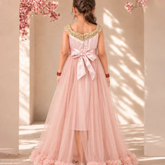 A young girl viewed from behind wearing a formal dusty pink tulle dress with gold beaded shoulder straps, a large satin bow at the back waist, and ruffled trim along the hem, standing in a sunlit architectural setting with pink blossoms.